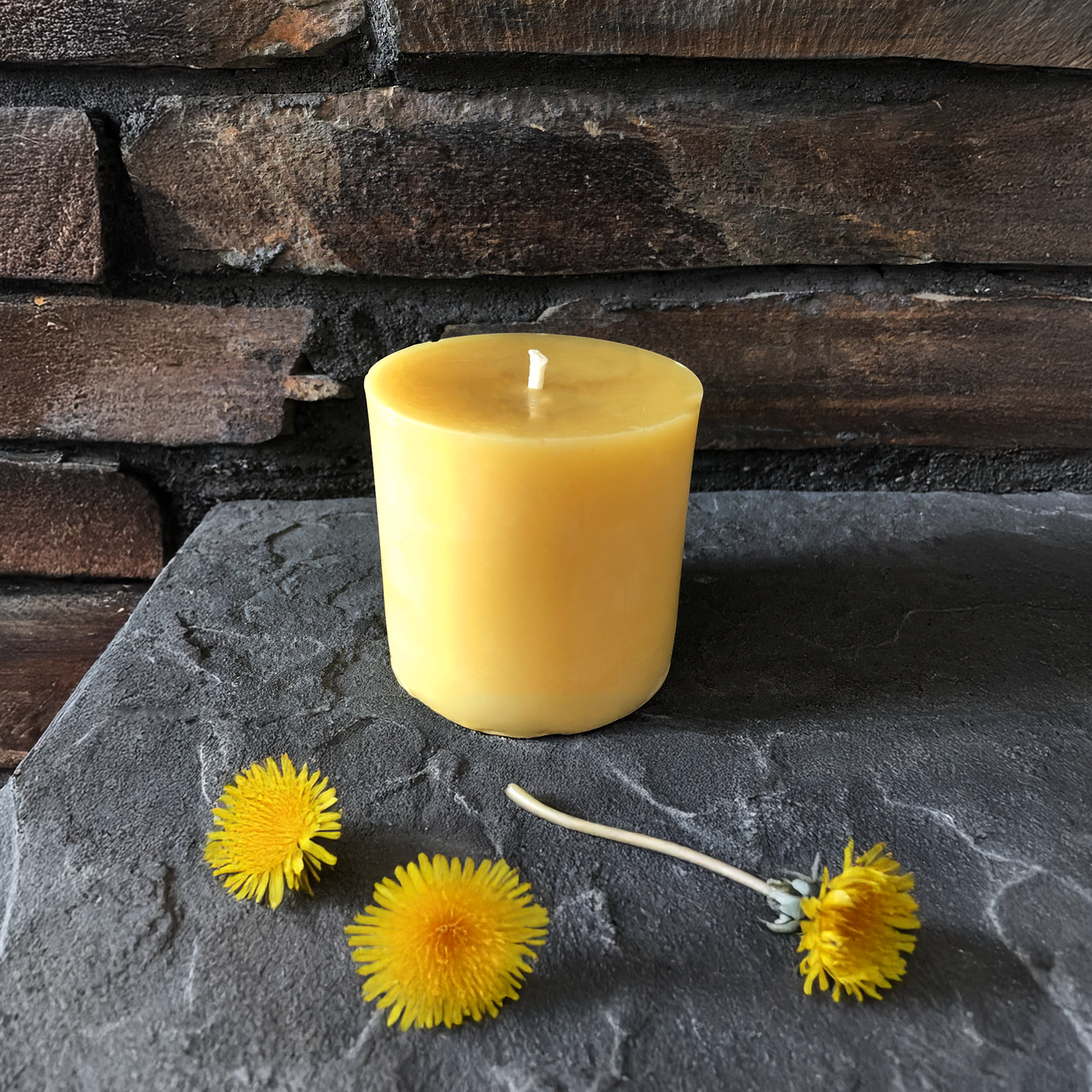 Yellow beeswax candle on a dark stone surface with dandelions against a stone wall background