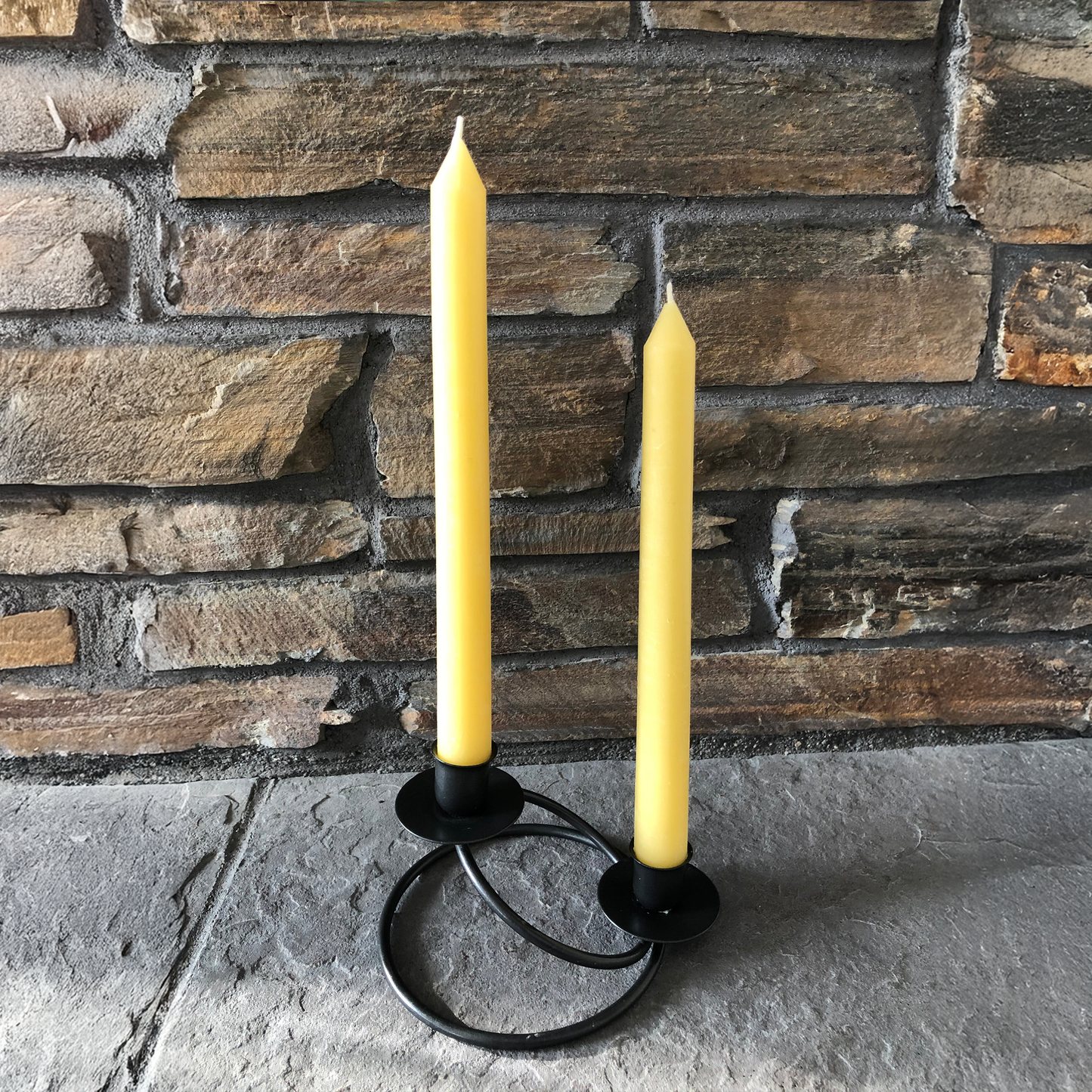 Two yellow beeswax candles in a black holder against a stone wall.