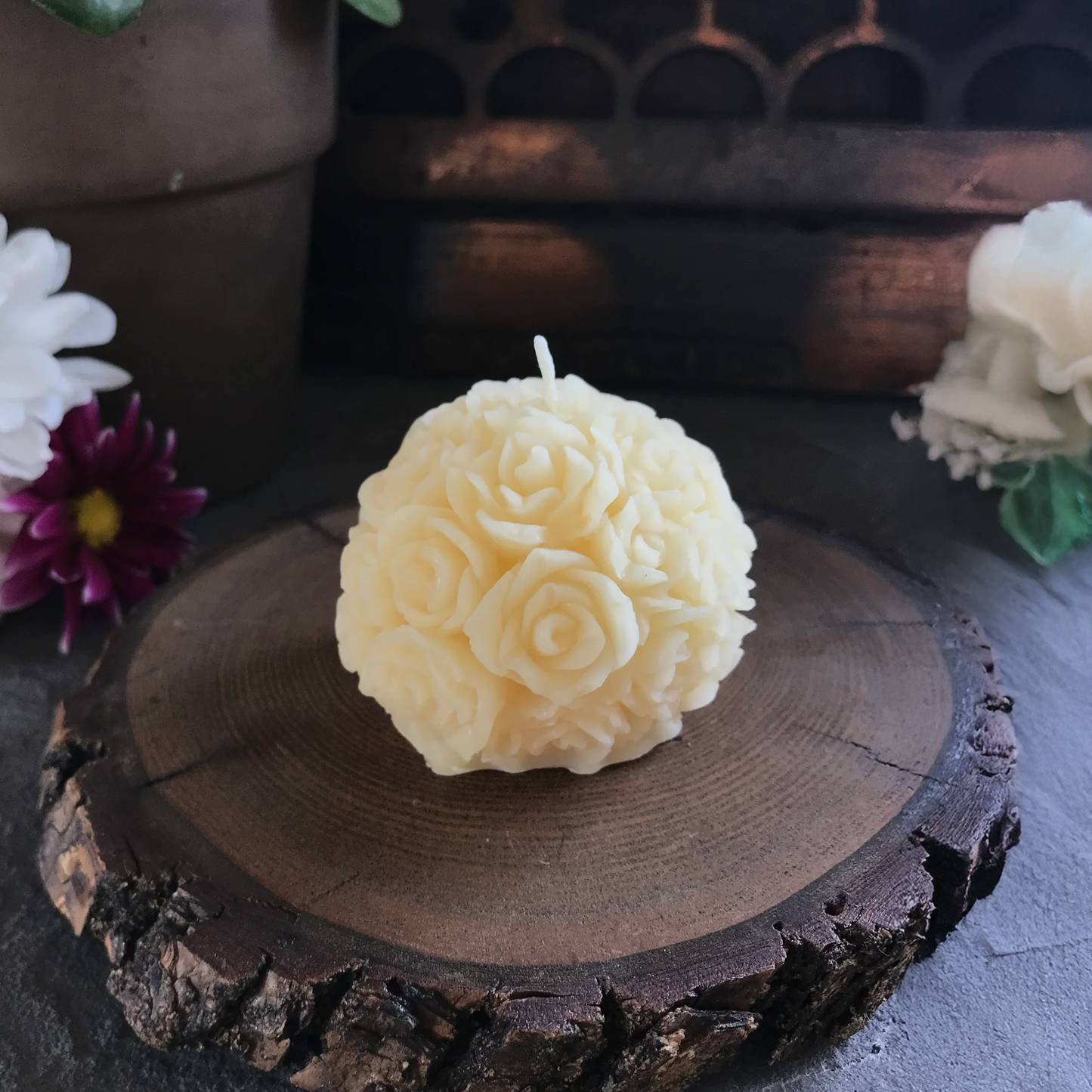 Decorative beeswax candle shaped like a flower ball on a wooden surface with flowers in the background
