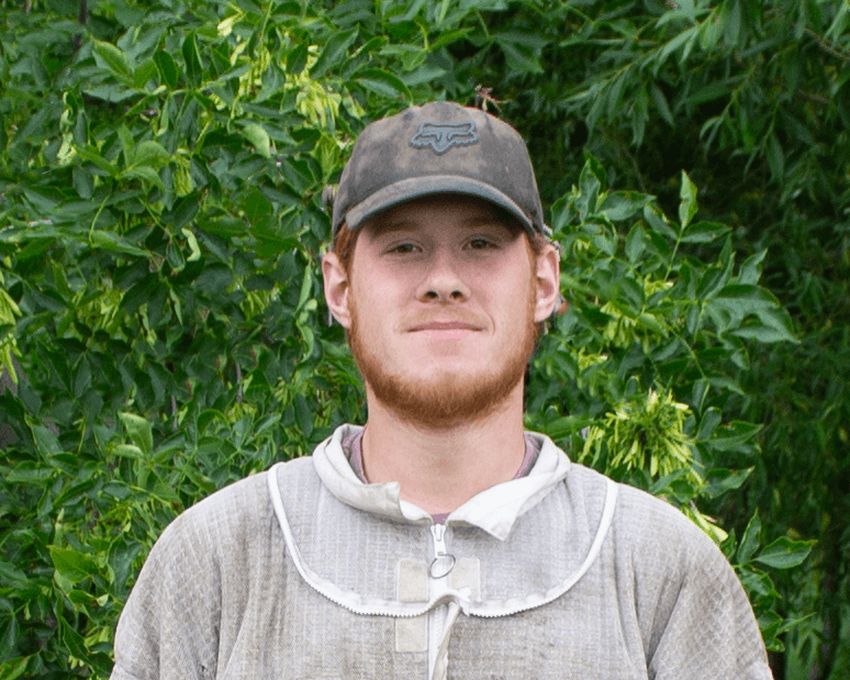 Man wearing a cap and beekeeper suit in front of green foliage