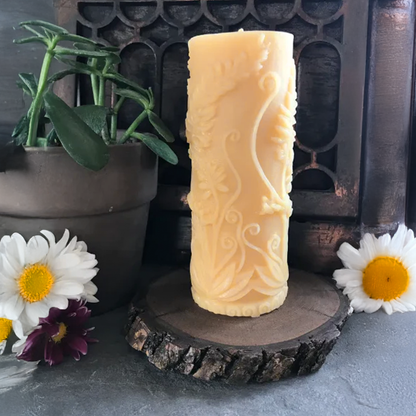 Decorative beeswax candle with intricate design on a wooden stand, surrounded by flowers and a pot.