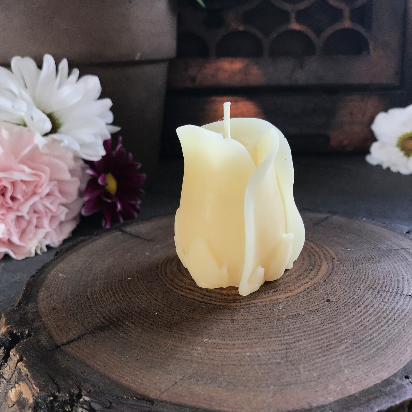White candle shaped like a rose on a wooden surface with flowers in the background