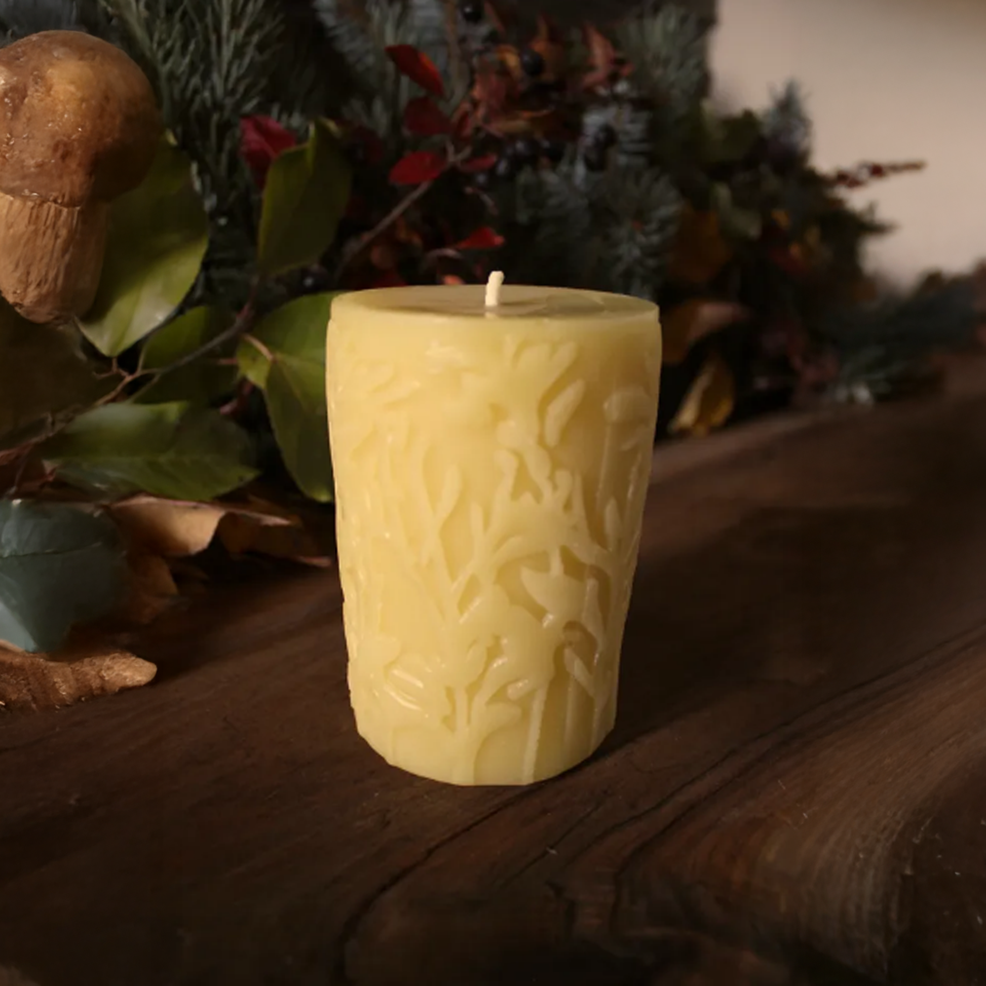 Decorative beeswax candle with leaf pattern on a wooden surface with plants in the background