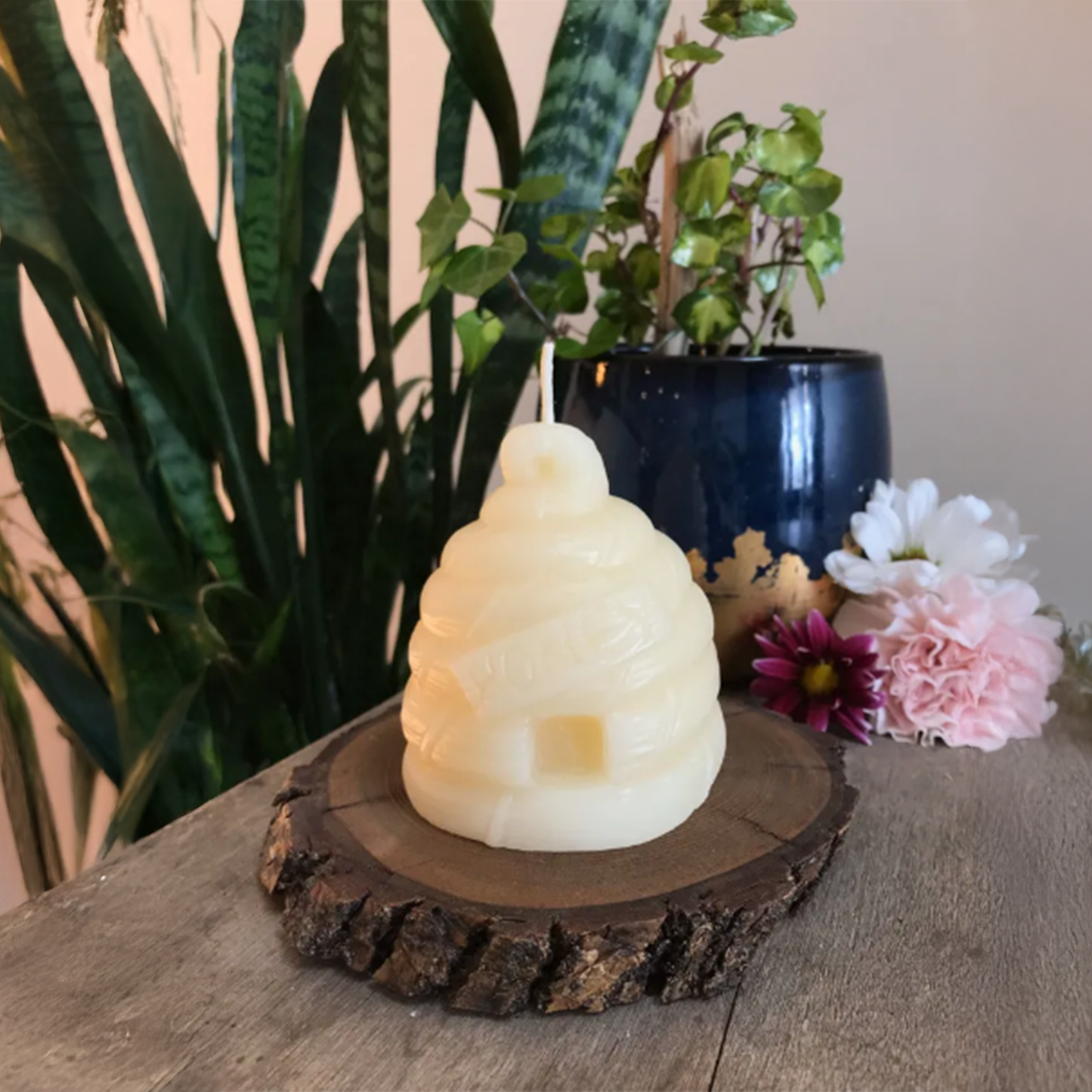 Beehive-shaped candle on a wooden base with plants and flowers in the background