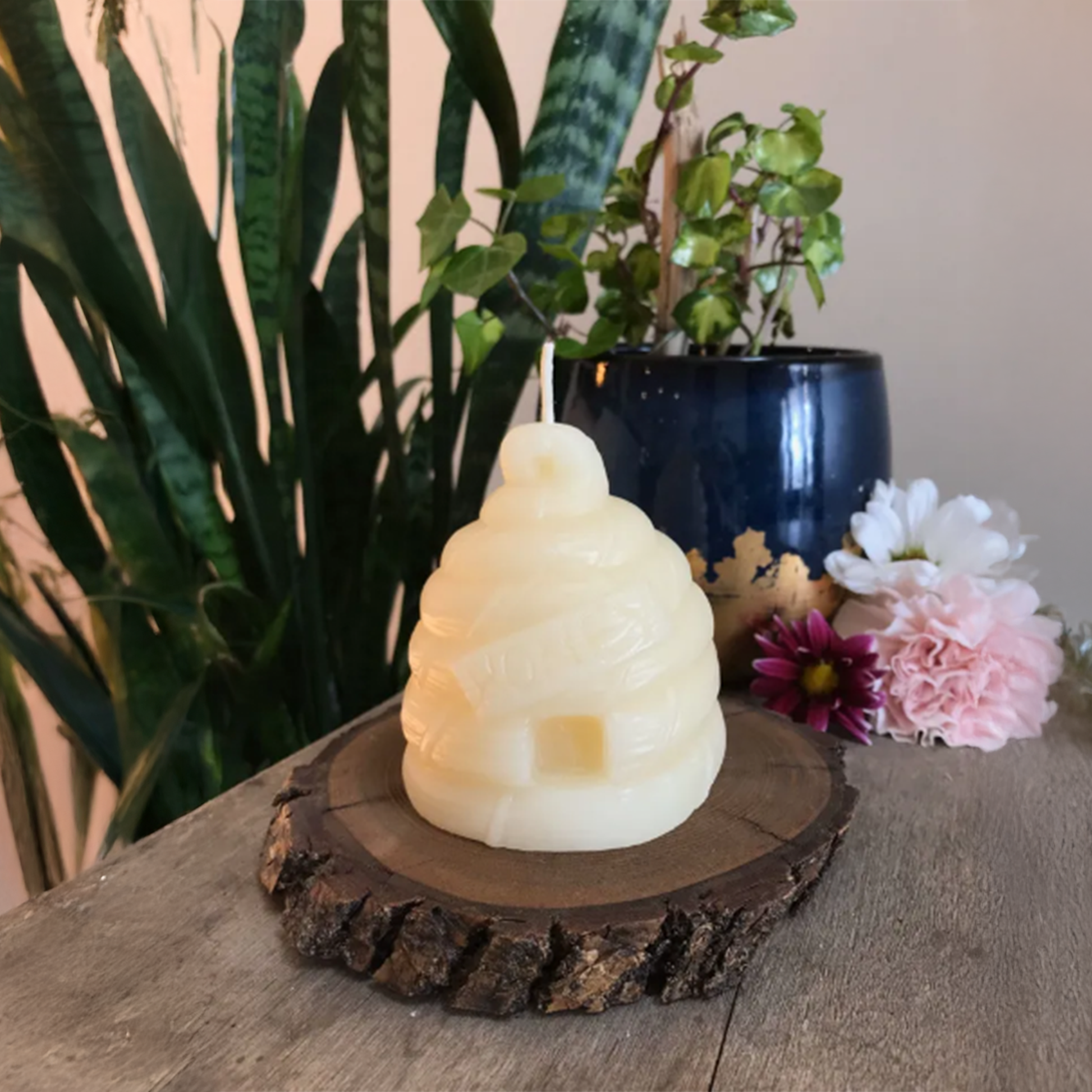 Beehive-shaped candle on a wooden base with plants and flowers in the background