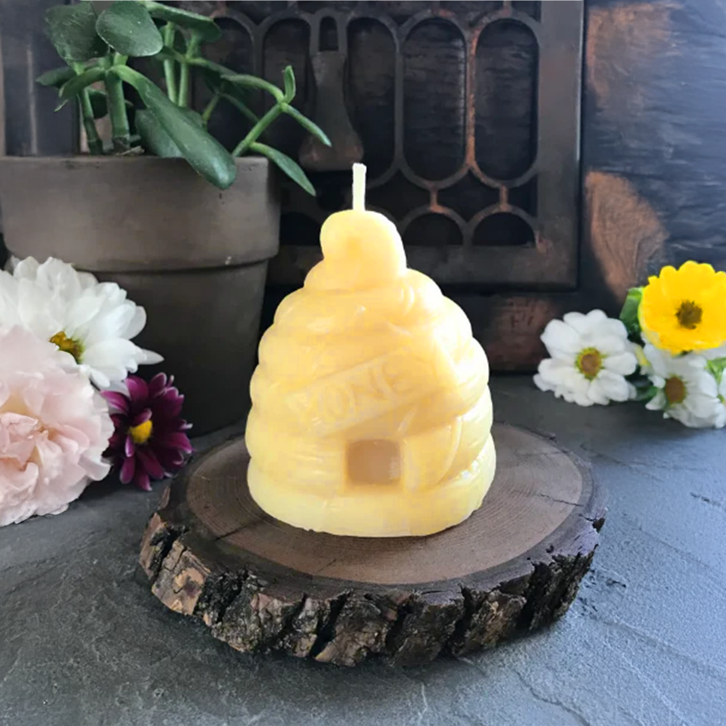 Beeswax candle shaped like a beehive on a wooden base with flowers and plants in the background.