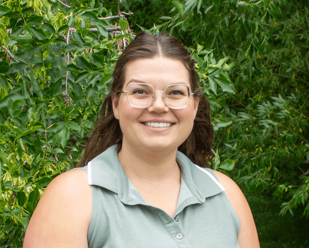 Woman wearing glasses and a light green sleeveless top standing in front of green foliage.