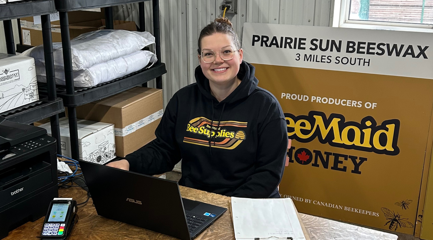 Person sitting at a desk with a laptop in front of a Prairie Sun Beeswax and BeeMaid Honey sign.