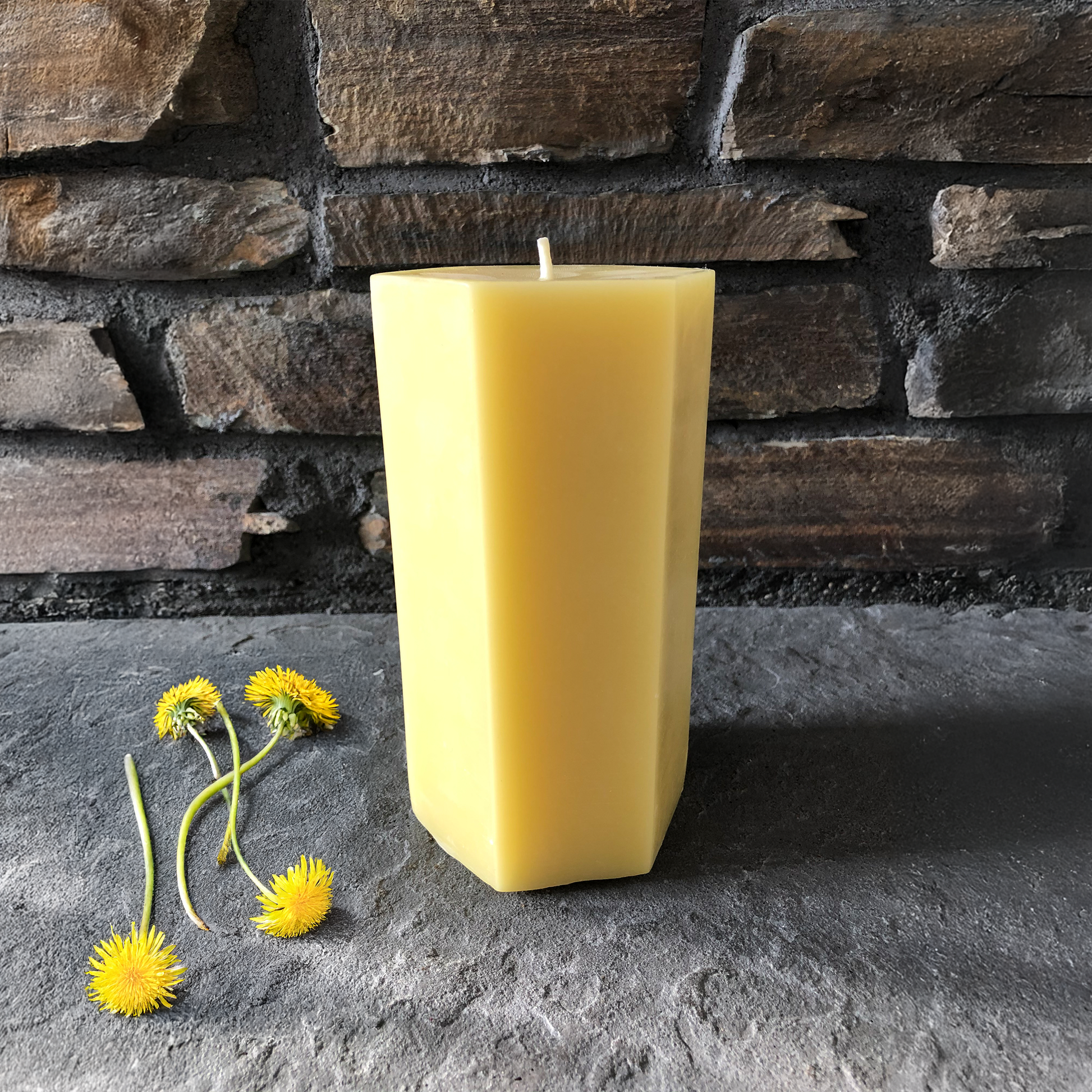 Yellow beeswax candle on a stone surface with a stone wall background and flowers