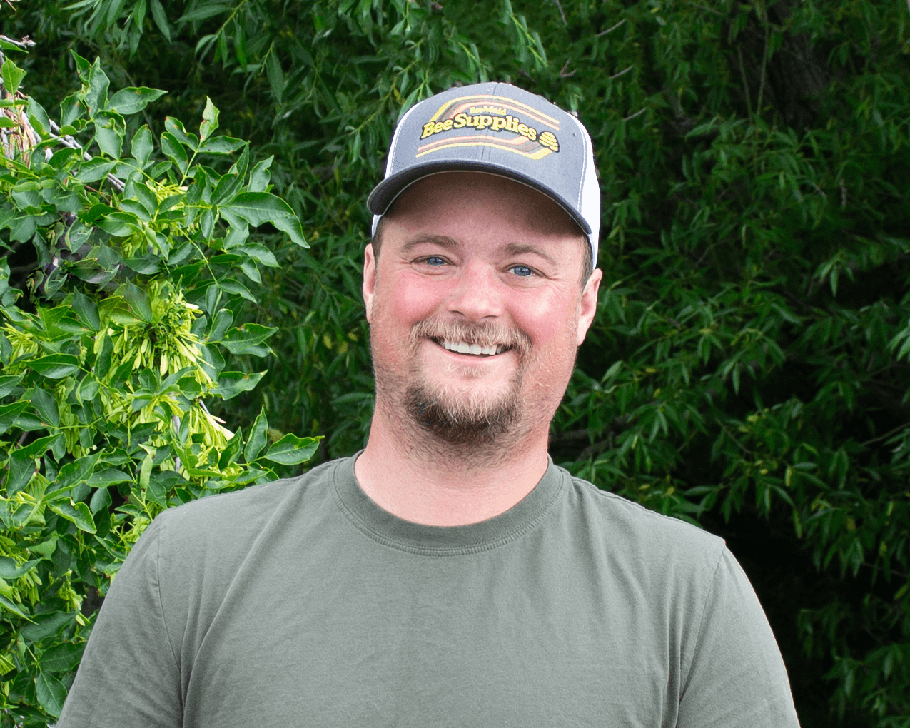 Man wearing a cap with 'Bee Supplies' logo in front of green foliage