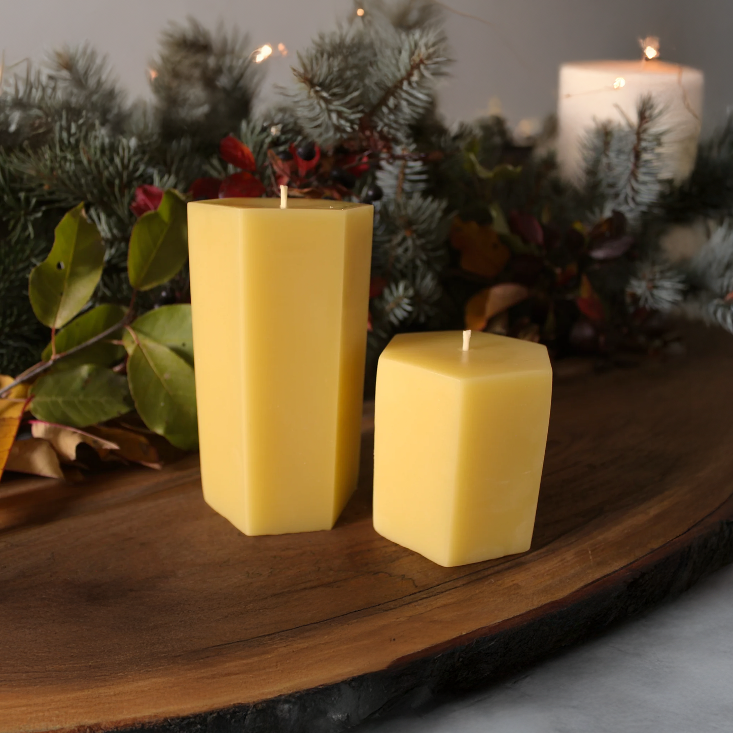 Two square yellow beeswax candles on a wooden surface with greenery in the background