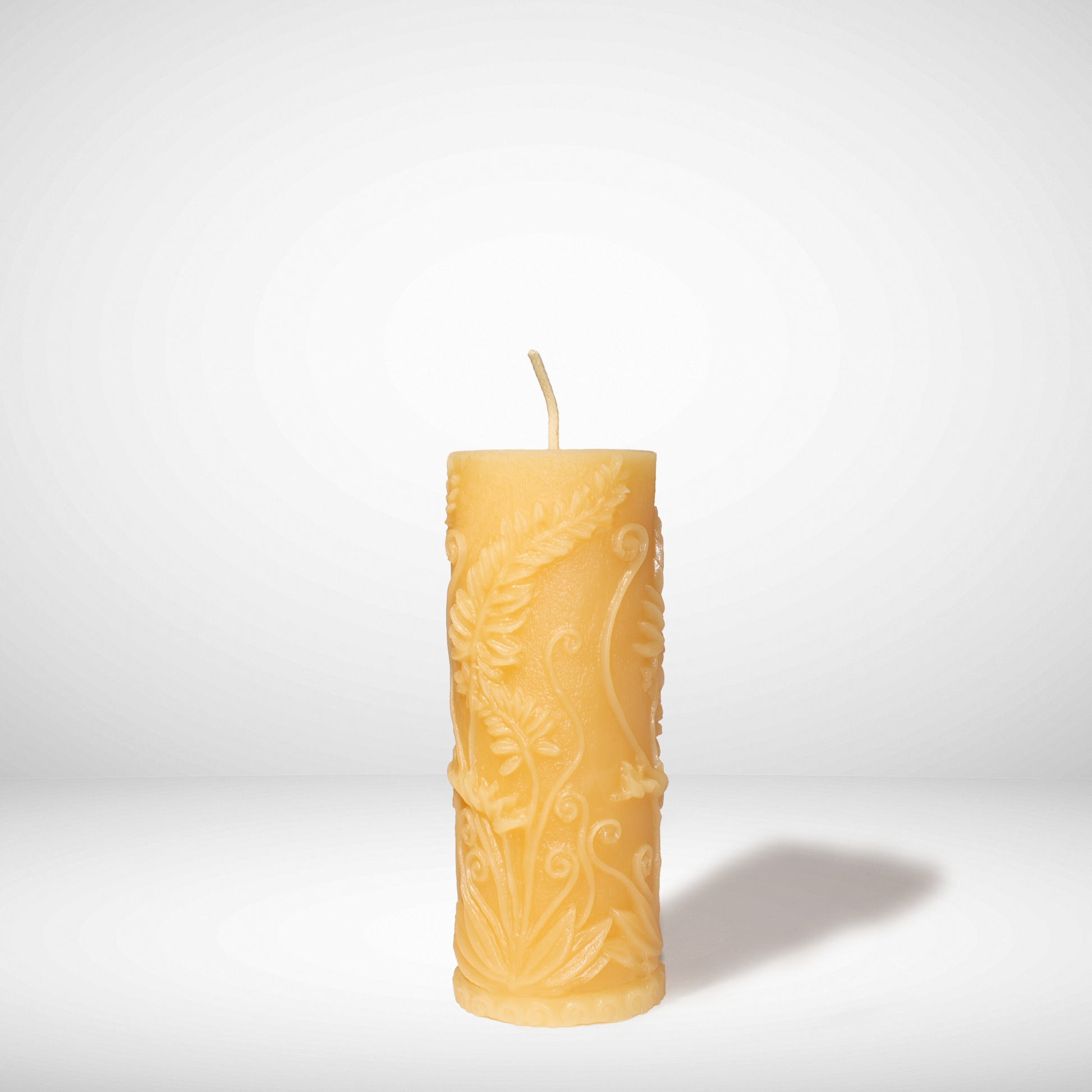 A Yellow beeswax candle with fern prints on the side in a pillar shaped, set against a white background. 