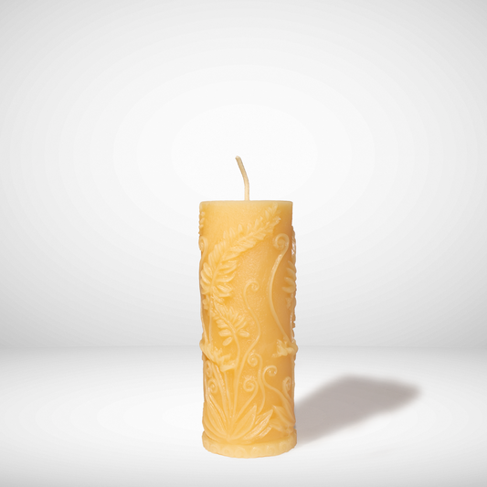 A Yellow beeswax candle with fern prints on the side in a pillar shaped, set against a white background. 