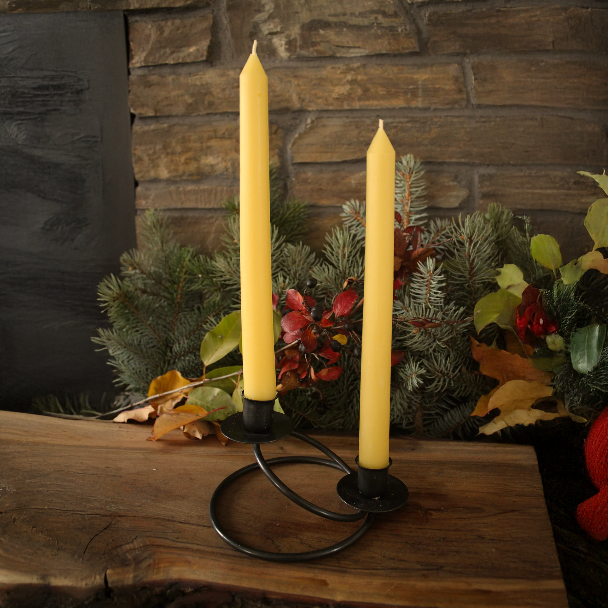 Two yellow beeswax candles on a black candlestick holder with a stone wall and foliage background.