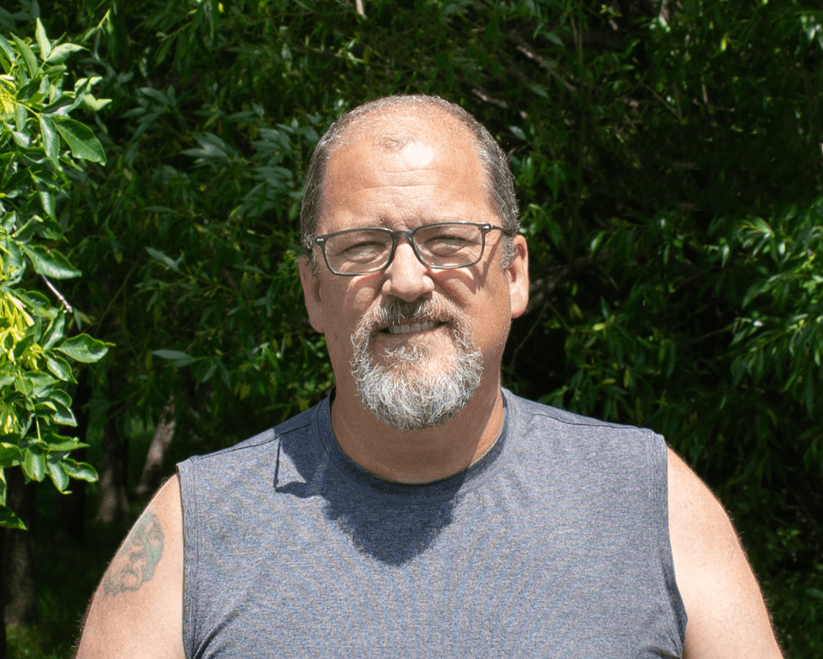 Man wearing glasses and a sleeveless shirt standing in front of green foliage